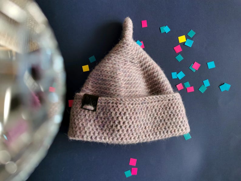 Op de afbeelding: Een licht mauve, getextureerde gebreide muts met een puntige bovenkant en een omgeslagen rand. Een klein, rechthoekig zwart label is bevestigd aan de linkerkant van de rand. De muts heeft een dicht, uniform steekpatroon en een zachte uitstraling.