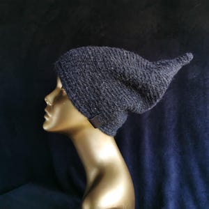 Op de afbeelding: Een donker antracietgrijze gebreide muts met een textuurrijke, gespikkelde uitstraling en een puntig, afhangend silhouet. De muts is voorzien van een klein, rechthoekig, bruin leerachtig label aan de onderrand van het ontwerp.