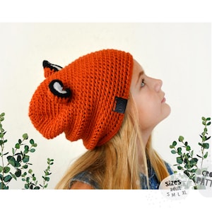 Puede incluir: Un gorro naranja tejido con características de zorro, incluyendo ojos y orejas en blanco y negro. El gorro tiene una apariencia texturizada y una pequeña etiqueta negra. La imagen también incluye el texto "Sizes Adult S M L XL" y "CROCHET PATTERN".