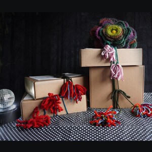 May include: An artistic display featuring stacked cardboard boxes adorned with colorful yarn embellishments. A large, multi-colored yarn flower sits atop the boxes, with trailing yarn accents. A disco ball adds a touch of sparkle to the scene.