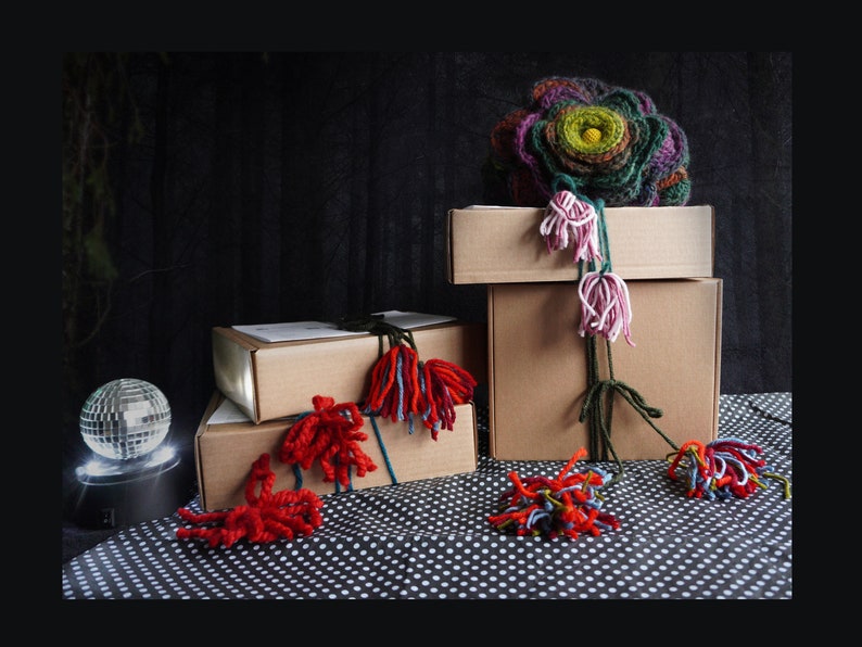 May include: An artistic display featuring stacked cardboard boxes adorned with colorful yarn embellishments. A large, multi-colored yarn flower sits atop the boxes, with trailing yarn accents. A disco ball adds a touch of sparkle to the scene.