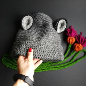 May include: A gray crocheted beanie featuring two rounded ears with light gray centers. Attached to the side are two long green crocheted stems, each topped with a textured orange base and vibrant magenta fringe petals resembling thistle flowers.
