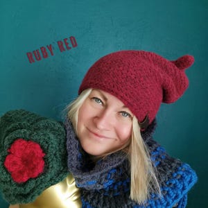 Op de afbeelding: Een robijnrode gehaakte muts met een klein pomponnetje, een donkergroene gehaakte hoed met een opvallend rood bloemaccent op een gouden etalagepophoofd, en een grove donkergrijze en blauwe gehaakte col met een franje aan de onderkant.