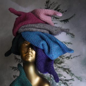 Op de afbeelding: Een goudkleurig etalagepophoofd met een stapel van meerdere gebreide mutsen in verschillende kleuren, waaronder lichtroze, magenta, grijs, blauw, paars en donkergroen. De mutsen zijn op elkaar gestapeld, wat zorgt voor een hoge, textuurrijke en kleurrijke hoop.