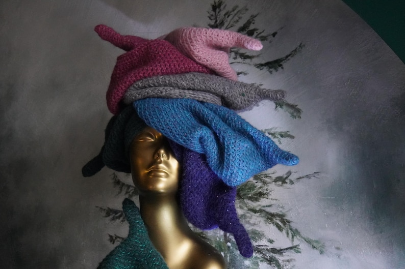 Op de afbeelding: Een goudkleurig etalagepophoofd met een stapel van meerdere gebreide mutsen in verschillende kleuren, waaronder lichtroze, magenta, grijs, blauw, paars en donkergroen. De mutsen zijn op elkaar gestapeld, wat zorgt voor een hoge, textuurrijke en kleurrijke hoop.