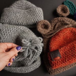 May include: A collection of handmade knitted hats in various colors and styles. The hats include a gray beanie, a gray hat with a ruffled edge, a brown hat with bear ears, and an orange hat with a leather label. The hats are made of a soft, textured yarn.