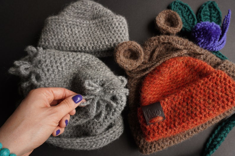 May include: A collection of handmade knitted hats in various colors and styles. The hats include a gray beanie, a gray hat with a ruffled edge, a brown hat with bear ears, and an orange hat with a leather label. The hats are made of a soft, textured yarn.