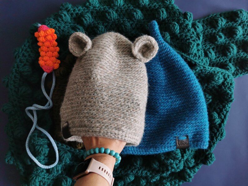 May include: A gray knit hat with attached ears and a blue knit hat are displayed on a textured green blanket. A small orange and pink carrot toy with a blue string is also present. The hats have a tag that says "Ulou".