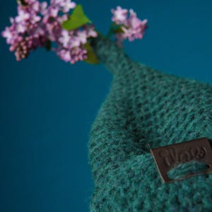 Op de afbeelding: Een donkergroene gebreide muts met een puntige bovenkant en een klein bruin rechthoekig leren label met de in reli&euml;f aangebrachte tekst "ilove". Een takje met lichtpaarse seringenbloemen steekt uit de bovenkant van de muts.