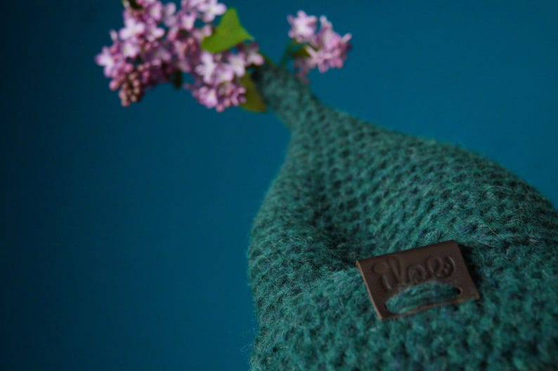 Op de afbeelding: Een donkergroene gebreide muts met een puntige bovenkant en een klein bruin rechthoekig leren label met de in reli&euml;f aangebrachte tekst "ilove". Een takje met lichtpaarse seringenbloemen steekt uit de bovenkant van de muts.