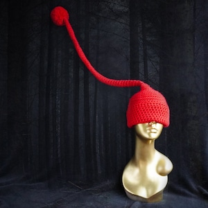 May include: A red crocheted hat with a long, pointed tail. The hat is on a mannequin head against a dark background.