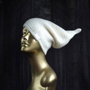 May include: A white crocheted beanie with a long, pointed tail. The beanie is on a mannequin head against a dark background.