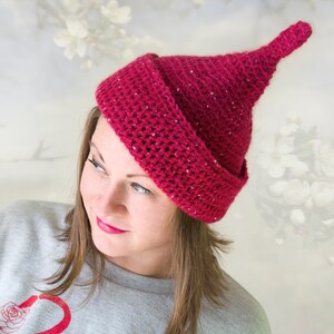 Op de afbeelding: Een diep magenta gehaakte muts met een puntige bovenkant en een brede, omgeslagen rand. Het garen bevat subtiele, ge&iuml;ntegreerde glitters door de hele textuur, wat een glinsterend effect cre&euml;ert over het oppervlak van de kegelvormige hoofdbedekking.