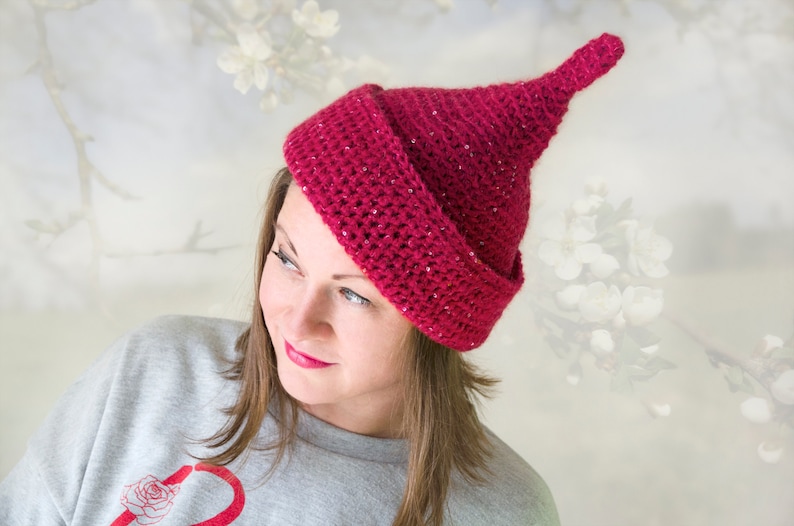Op de afbeelding: Een diep magenta gehaakte muts met een puntige bovenkant en een brede, omgeslagen rand. Het garen bevat subtiele, ge&iuml;ntegreerde glitters door de hele textuur, wat een glinsterend effect cre&euml;ert over het oppervlak van de kegelvormige hoofdbedekking.