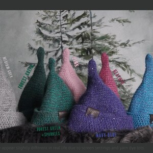 Op de afbeelding: Een collectie van zeven gehaakte puntmutsen in diverse kleuren, waaronder middengrijs, bosgroen, bosgroen met glitters, roze, paars, heidekleur en pauwblauw. Elke muts is voorzien van een omgeslagen rand en een klein bruin leren label.