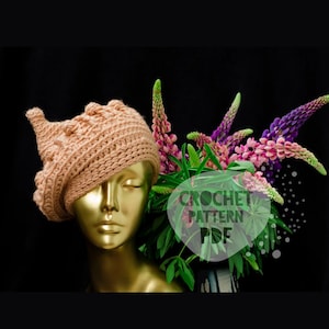 Op de afbeelding: Een beige gehaakte hoed met een uniek ontwerp wordt getoond op een gouden mannequinhoofd. Een boeket paarse en roze bloemen staat naast de hoed. De afbeelding bevat de tekst "CROCHET PATTERN PDF".