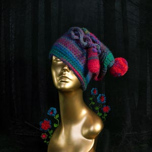 Pode incluir: Um chapéu colorido, tricotado à mão, com um design extravagante, apresentando tons de azul, roxo e verde, acentuado com pompons vermelhos e elementos pendurados. O chapéu é exibido em uma cabeça de manequim dourada.