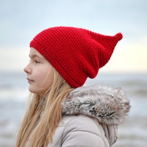 May include: A young person wearing a red knitted beanie with a long pointed tip. The beanie is pulled down over their ears and forehead. The person is looking off to the side, with a thoughtful expression on their face. They are standing in front of a body of water, with waves crashing in the background.