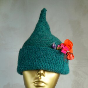 Op de afbeelding: Een donkergroene, puntige gehaakte muts met een brede, omgeslagen rand. De muts is versierd met een klein boeketje van feloranje, roze en paarse bloemen die aan de zijkant van de rand zijn ingestoken, wat kleur toevoegt aan het textuurrijke garen.