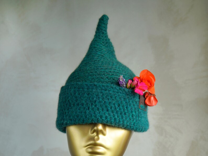 Op de afbeelding: Een donkergroene, puntige gehaakte muts met een brede, omgeslagen rand. De muts is versierd met een klein boeketje van feloranje, roze en paarse bloemen die aan de zijkant van de rand zijn ingestoken, wat kleur toevoegt aan het textuurrijke garen.