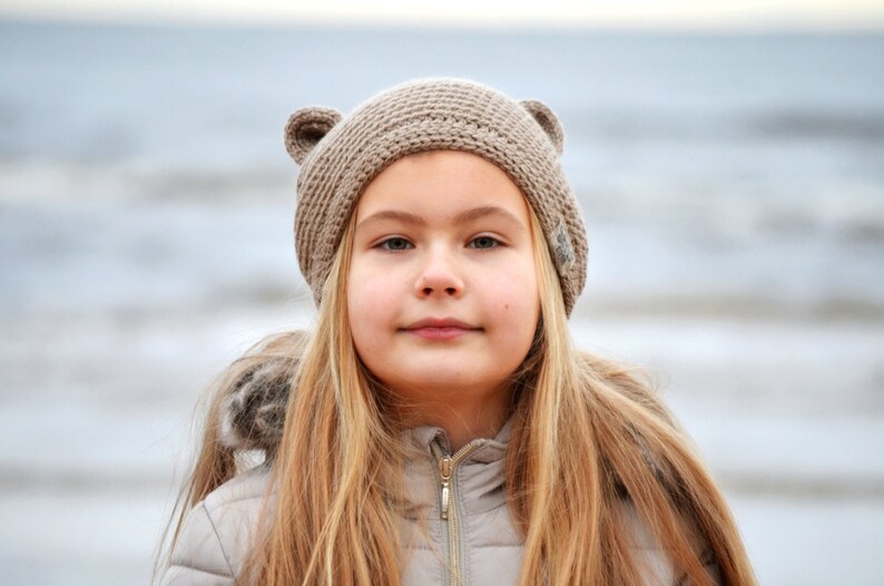 May include: A young person with long blonde hair wearing a tan knit hat with bear ears. The person is also wearing a light-colored jacket with a fur-lined hood. The background is a blurred view of the ocean.