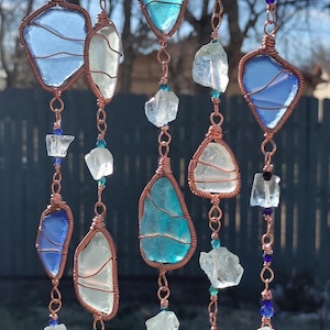 May include: A handcrafted wind chime featuring sea glass and clear crystal accents. The sea glass pieces are in shades of blue and white, wrapped in copper wire, and strung together with beads and chains. A decorative outdoor accent.