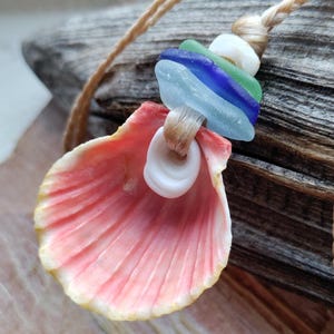 May include: A handcrafted necklace featuring a pink seashell pendant with a cream-colored rim. The necklace includes sea glass beads in blue, green, and white, strung on a natural fiber cord. The seashell is approximately 3 inches wide.