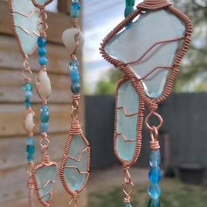 May include: Handmade wind chimes featuring sea glass in shades of blue and turquoise, wrapped in copper wire. Accented with blue beads and small seashells, these chimes add a coastal touch to any space.