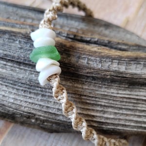 May include: A macrame bracelet crafted from tan cord featuring a central arrangement of three white shell beads and one green sea glass bead. The bracelet is constructed with a twisted knot pattern and includes a small wooden bead at the closure.