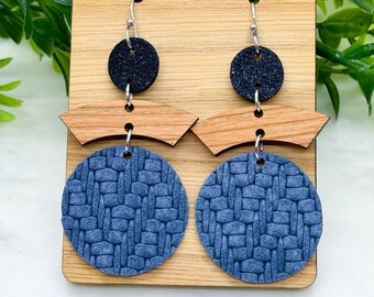 May include: A pair of dangle earrings with a woven blue leather circle and a smaller black glitter circle. The earrings are hanging from a wooden display card with the text "DaisyLove Studios".