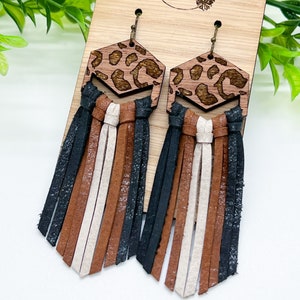 May include: Leopard print wooden earrings with black, brown, and cream leather fringe. The earrings are on a wooden display card with the text "Daisy Love Studios".