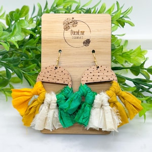 May include: A pair of handmade earrings with a wooden half-circle top featuring a dotted pattern. The earrings have gold-colored hooks and are adorned with fabric tassels in yellow, green, and off-white. The wooden display has the text "DaisyLove Studios".
