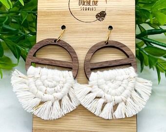 May include: A pair of wooden earrings with a white macrame tassel and gold accents. The earrings are displayed on a wooden card with the text "DaisyLove Studios".