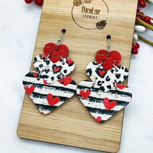 May include: A pair of dangle earrings featuring three layered heart shapes. The hearts are made of cork and feature a red, black, and white striped pattern, a leopard print, and a solid red pattern. The earrings are hanging from silver hooks.
