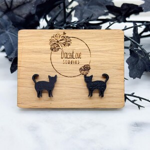 May include: A pair of black cat silhouette earrings on a wooden display card with the text "DaisyLove Studios".
