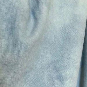 May include: Close-up of a light blue fabric with a textured appearance. The fabric has a soft, slightly faded look, with subtle variations in color and tone. The material appears to be lightweight and possibly cotton or a similar blend.