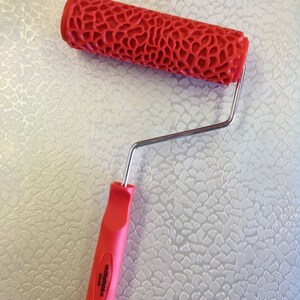 May include: Red paint roller with a textured pattern and a silver handle. The roller is designed to create a textured finish on walls or other surfaces.