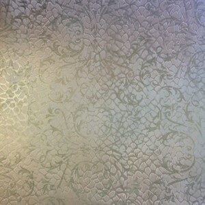 May include: A textured, light beige background with a subtle, repeating pattern of swirling vines. The pattern is embossed and creates a raised, three-dimensional effect.