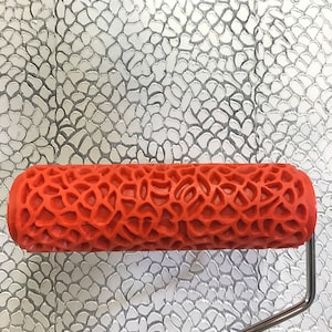 May include: A red paint roller with a textured pattern of small, irregular shapes. The roller is designed to create a decorative, textured finish on walls or other surfaces.