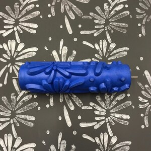 May include: Blue paint roller with a pattern of fireworks. The roller is on a dark gray surface with a white firework pattern.