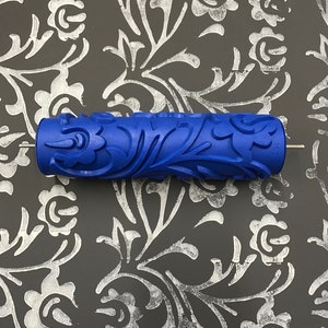May include: A blue paint roller with a floral design. The roller is on a dark gray surface with a white floral pattern.