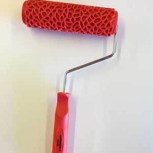 May include: Red paint roller with a textured pattern and a silver handle. The handle has a red plastic grip with the text "wooddesigns.com" printed on it.