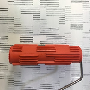 May include: An orange paint roller with a textured design is positioned in front of a wall with a repeating pattern of horizontal lines. The roller has a metal handle and is ready for use.