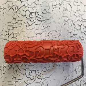 May include: An orange textured paint roller with a geometric pattern. The roller is made of a rubber-like material and has a silver metal handle. The background shows a sample of the pattern the roller creates.