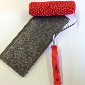 May include: A red paint roller with a textured pattern, designed to create a stone-like effect. The roller is attached to a metal handle with a red plastic grip. The handle has the text "Made in USA" printed on it.