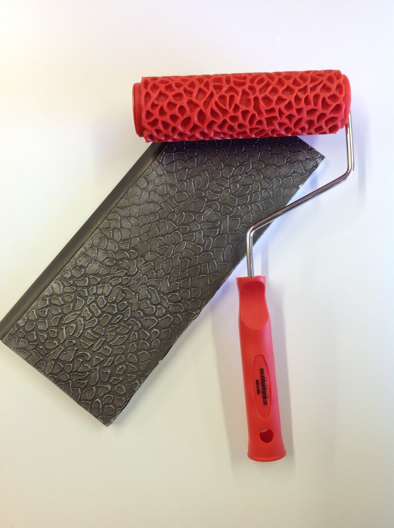 May include: A red paint roller with a textured pattern, designed to create a stone-like effect. The roller is attached to a metal handle with a red plastic grip. The handle has the text "Made in USA" printed on it.