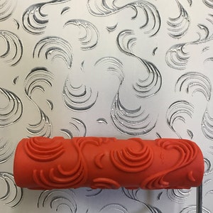 May include: An orange paint roller with a decorative floral pattern is positioned over a wall with a matching embossed design. The roller's handle is silver, and the wall's background is a light gray.