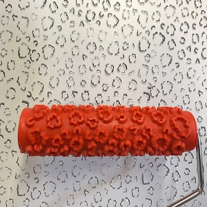 May include: A red paint roller with a pattern of small flowers. The roller is on a white surface with a repeating pattern of small, irregular shapes.