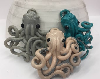 Op de afbeelding: Drie handgemaakte octopus-hangers in grijstinten, blauwgroen en beige. Elke octopus heeft zwarte ogen en gedetailleerde tentakels. De hangers zijn waarschijnlijk gemaakt van polymeerklei of een vergelijkbaar materiaal, met een grillig ontwerp met oceaanthema.