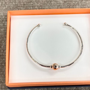 Könnte beinhalten: Ein silbernes Armreif-Armband mit einem goldenen Kugel-Anhänger in einer weißen Box mit orangefarbenem Deckel.